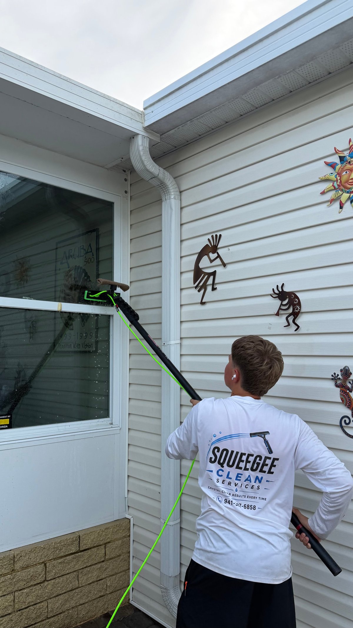 Worker from Squeegee Clean Services using a pole squeegee to clean siding and window on a light blue house with tribal artwork